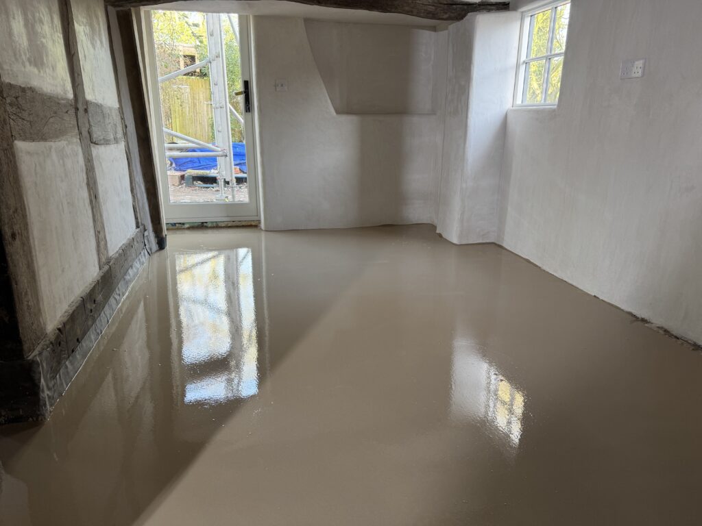 A freshly poured, smooth, and glossy self-leveling floor inside a room with white plaster walls, exposed wooden beams, and a glass door leading outside. Natural light reflects off the wet surface.