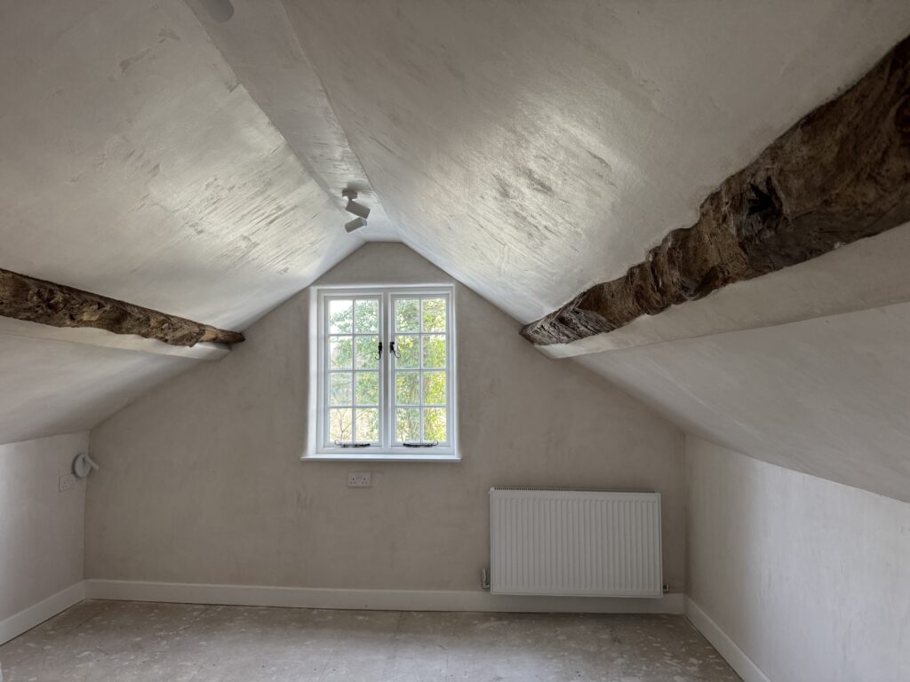 An empty attic room with slanted white walls, exposed wooden beams, a window with divided panes letting in natural light, and a radiator beneath the window. The floor appears unfinished.
