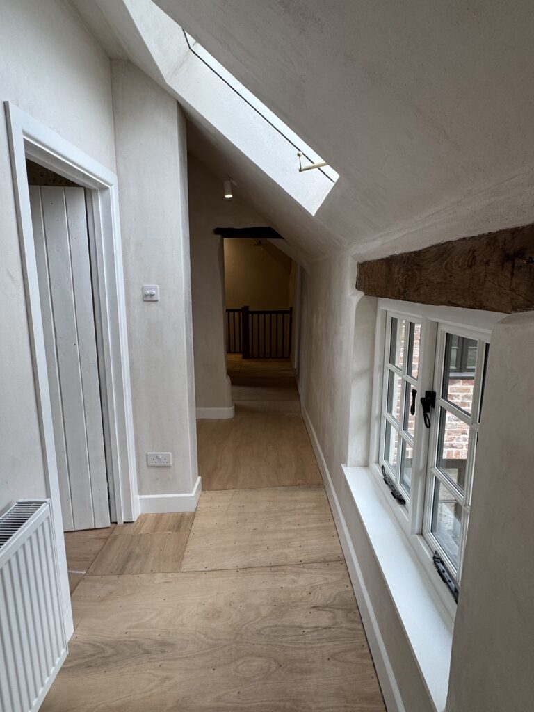 A narrow hallway with light wooden floors, white walls, a skylight, a rustic wooden beam above a window on the right, and a door on the left. The hallway leads to an open area with a black railing.
