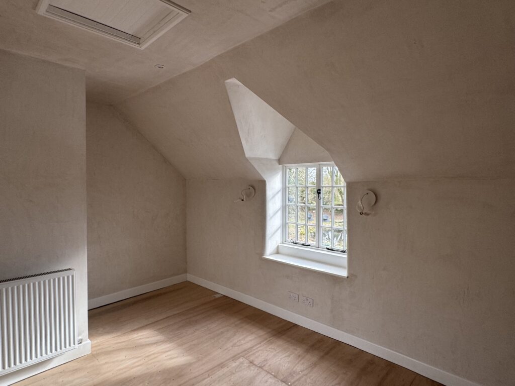 An empty room with cream-colored walls, a slanted ceiling, a white radiator, wooden floors, and a window with divided panes letting in natural light. Two wall fixtures are mounted on either side of the window.