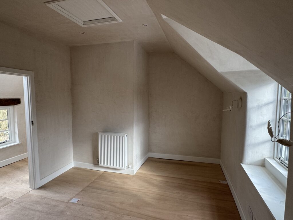 Small empty room with beige walls, light wood flooring, a radiator on one wall, a slanted ceiling, and a large window letting in natural light. An open door leads to an adjacent room.
