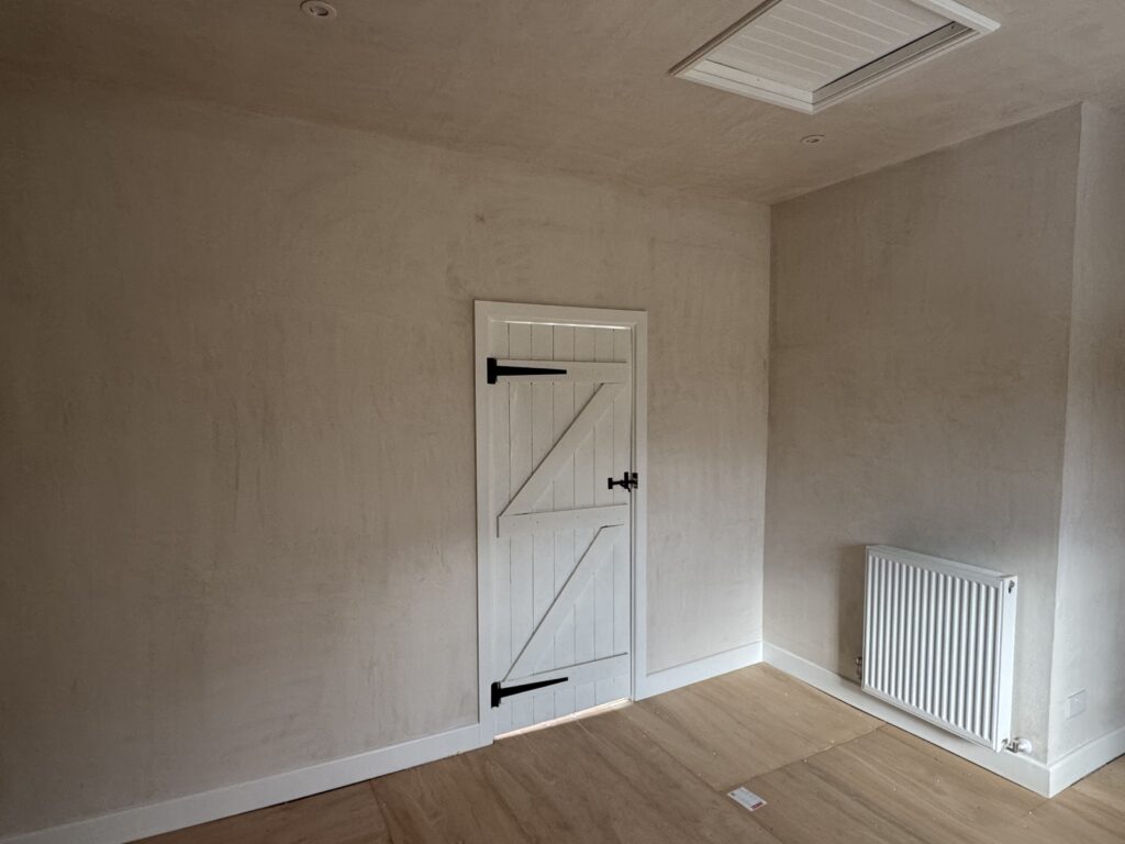 A small white wooden door with black hinges and latch is set in a plain, beige wall. A white radiator is mounted on the right, and light wood flooring covers the room. The ceiling has a rectangular hatch.