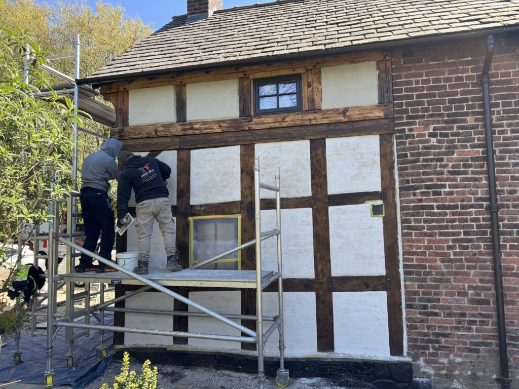 Two people stand on scaffolding while painting the exterior of a half-timbered house with white paint. The house has exposed wooden beams and red brick, and trees are visible in the background.