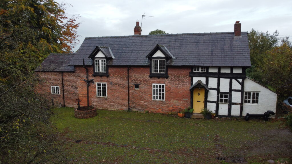 A two-story brick and timber-framed cottage with a gray slate roof, black-and-white accents, green grass lawn, potted plants by the door, and trees in the background on a cloudy day.