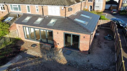 Aerial view of a brick house extension with large glass doors, skylights on the roof, and a partially finished yard with exposed soil and gravel. Fence and neighboring houses are visible.