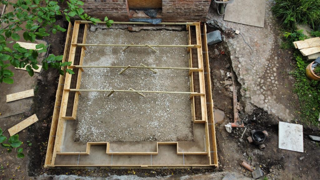 Aerial view of a rectangular construction site with wooden framing and gravel base, prepared for a foundation next to a brick building. Tools, wood planks, and construction materials are scattered around the site.