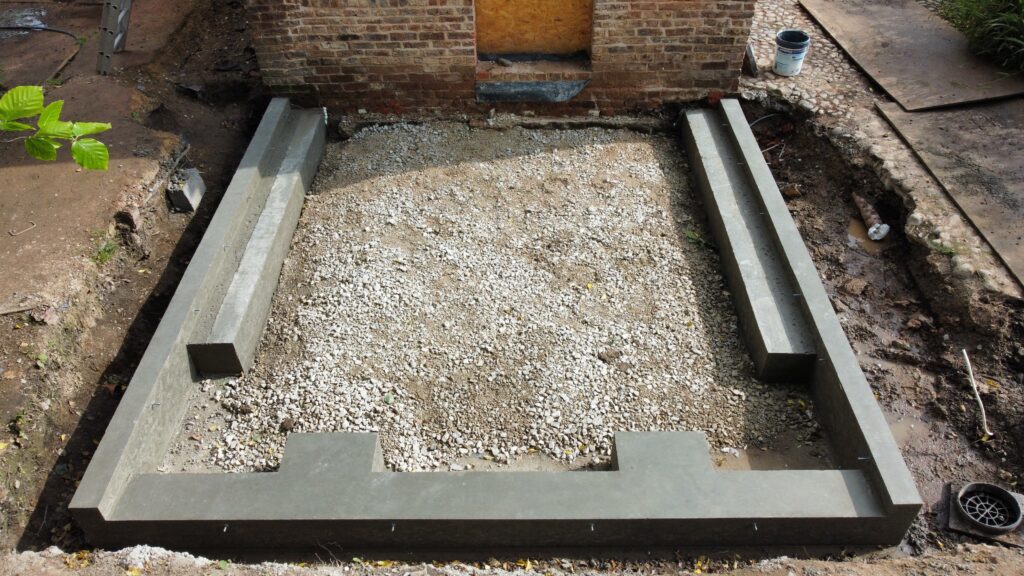 Concrete foundation footings form a rectangular shape with a gravel base inside, situated next to a brick wall. Construction materials and soil are visible around the foundation area.