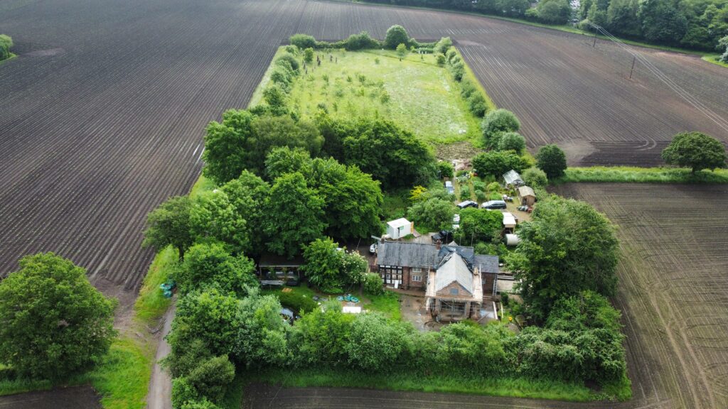 Aerial view of a farmhouse surrounded by dense trees, gardens, and fields of tilled soil. The property is bordered by greenery, with a large open grassy area behind the house and a dirt road on one side.