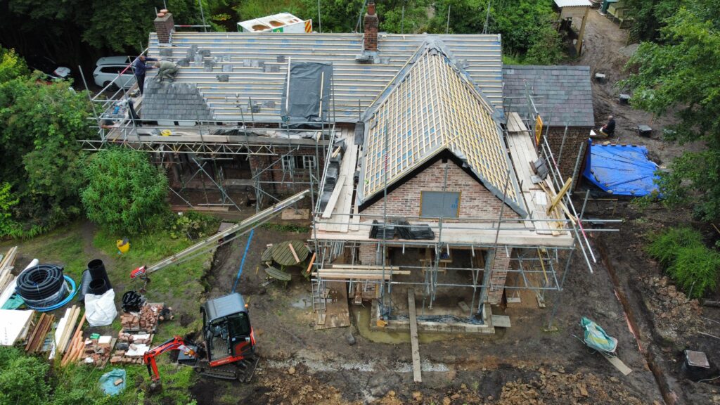 Aerial view of a house under renovation with scaffolding around the structure, roof work in progress, construction materials scattered outside, and a small excavator on muddy ground nearby.
