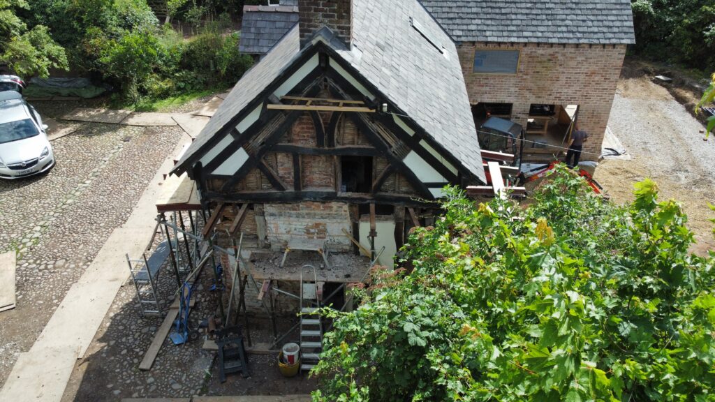 Aerial view of a half-timbered house under renovation, with exposed wooden beams and scaffolding at the front; tools and construction materials are scattered around the site, and a car is parked nearby.