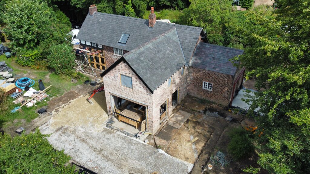 Aerial view of a house with an unfinished brick extension. The construction area is surrounded by building materials, tools, and cleared land, with trees and greenery around the property.