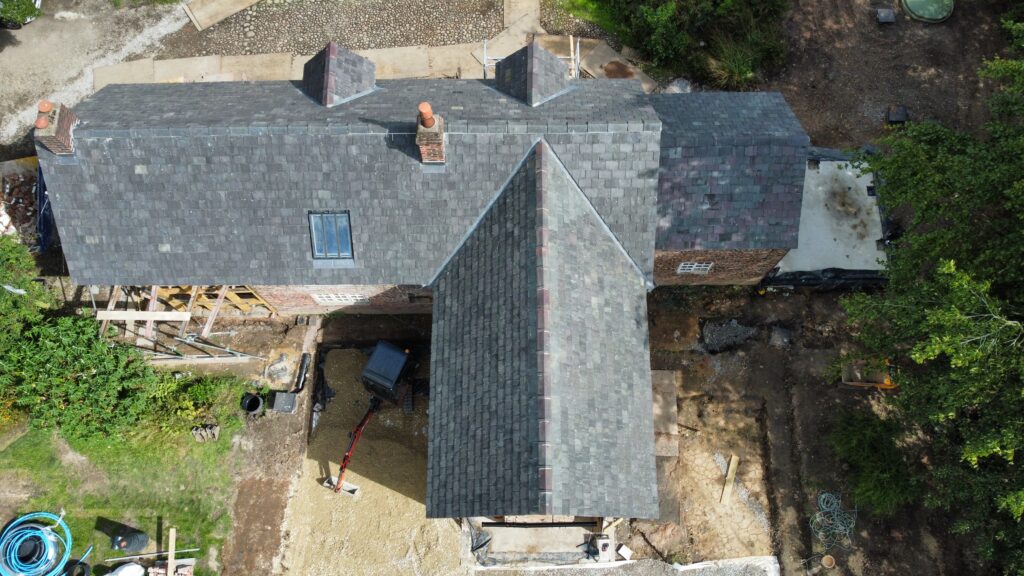 Aerial view of a house with a dark gray, pitched roof and two chimneys, surrounded by construction materials and equipment, with areas of exposed ground and some greenery at the edges.