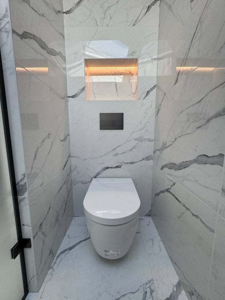 A modern bathroom with white marble walls and floor, a wall-mounted toilet, a rectangular niche with warm lighting above it, and a glass door partially visible on the left.