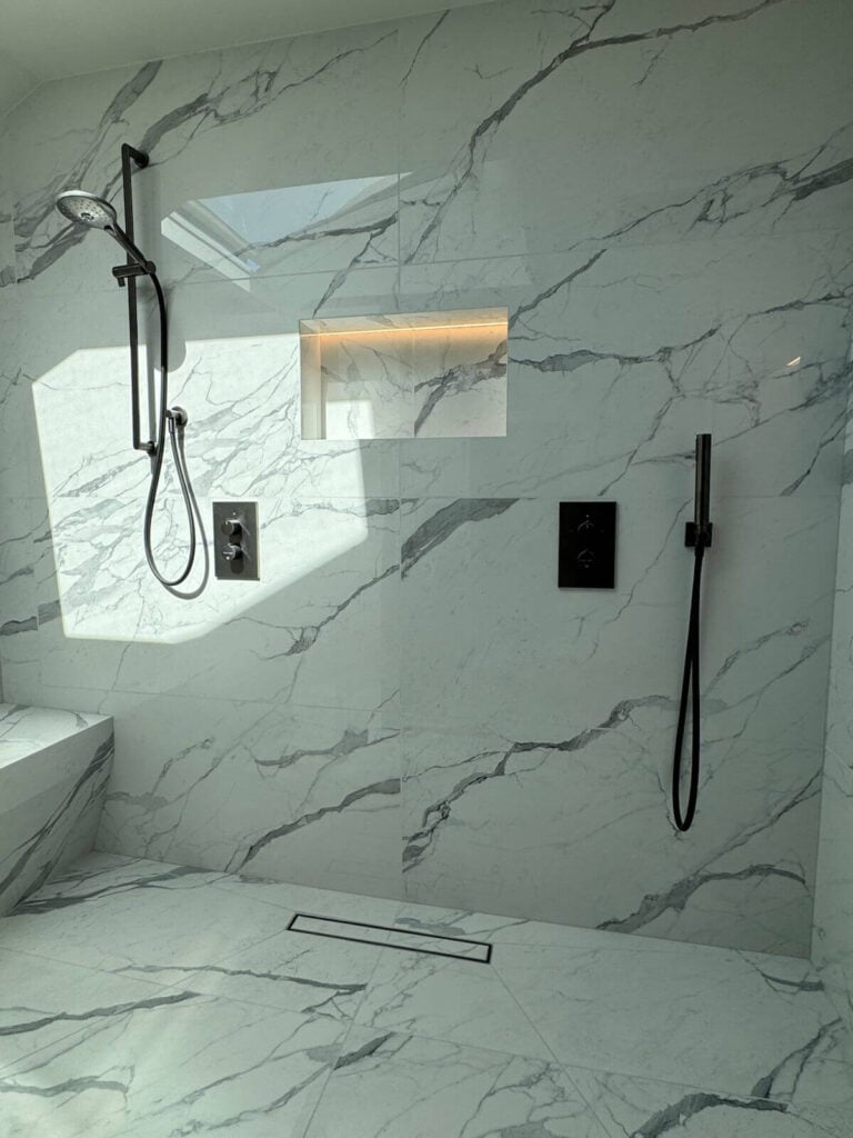 A modern shower with white marble walls, a built-in shelf, a handheld showerhead on the left, a wall-mounted control panel, and a sleek linear drain on the floor. Natural light streams in from above.