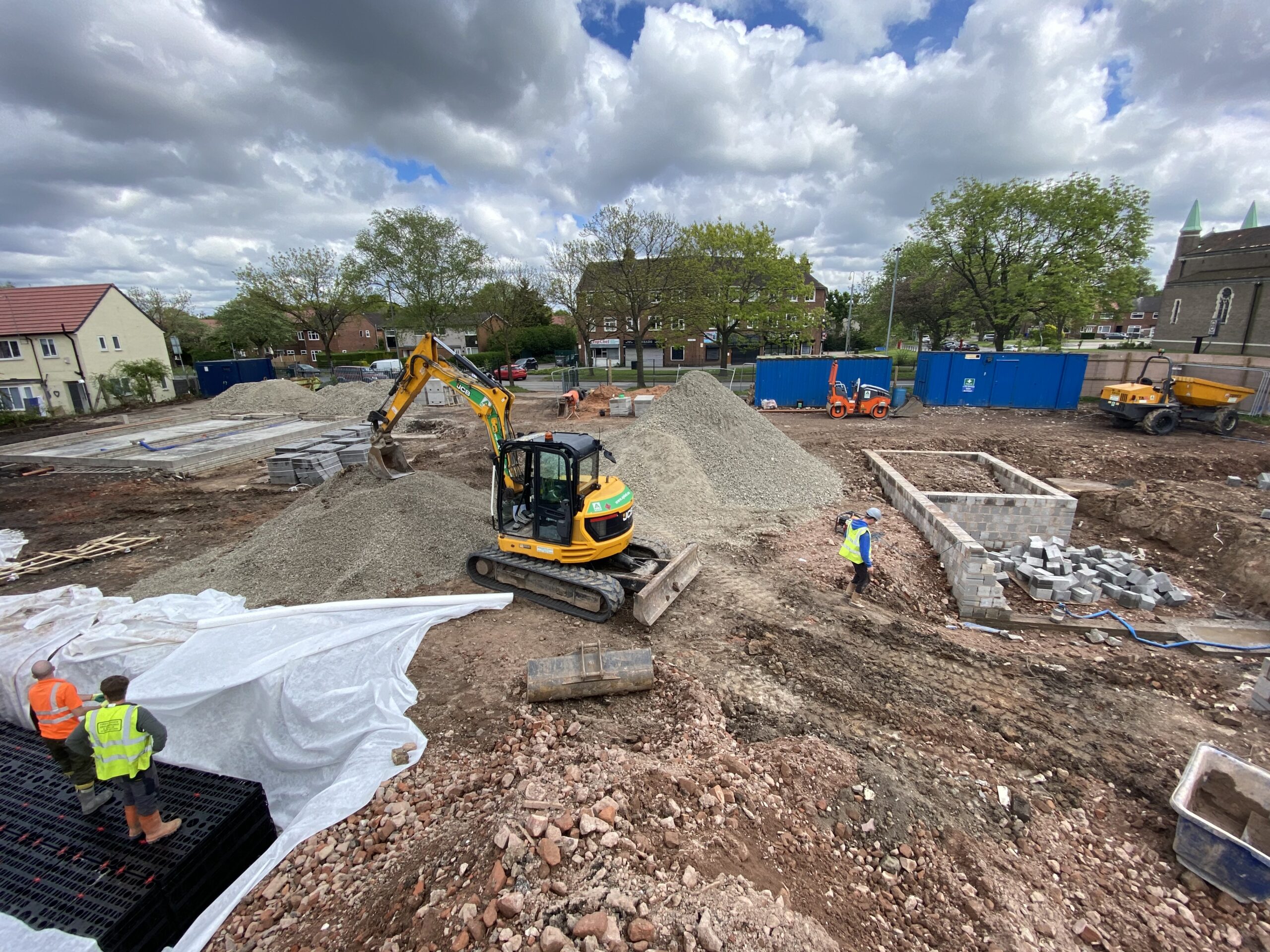 A construction site with workers in safety vests, a small yellow excavator moving gravel, piles of building materials, and unfinished structures surrounded by trees and blue temporary fencing under a partly cloudy sky.