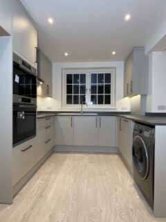 Modern kitchen with light gray cabinets, built-in oven and microwave on the left, washing machine on the right, central sink under a window, under-cabinet lighting, and light wood flooring.