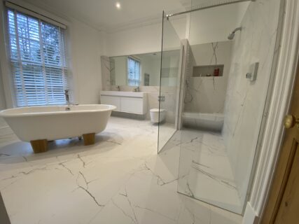 A modern bathroom with white marble floors and walls, a freestanding bathtub, a large mirror, a glass-enclosed shower, and a wide window with white blinds.