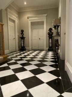 Elegant hallway with black and white checkered marble floor, two white doors, classical busts on pedestals, a table with a lamp and vase, and wooden staircase on the left.