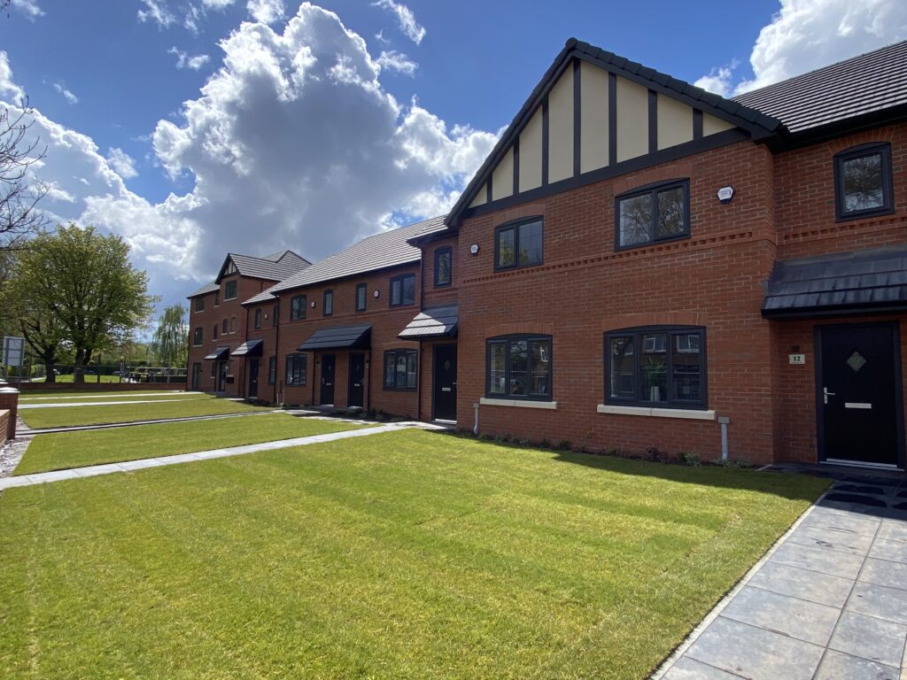 A row of modern brick townhouses with black roofs and large windows, set behind neatly mowed lawns and a stone pathway under a partly cloudy blue sky.