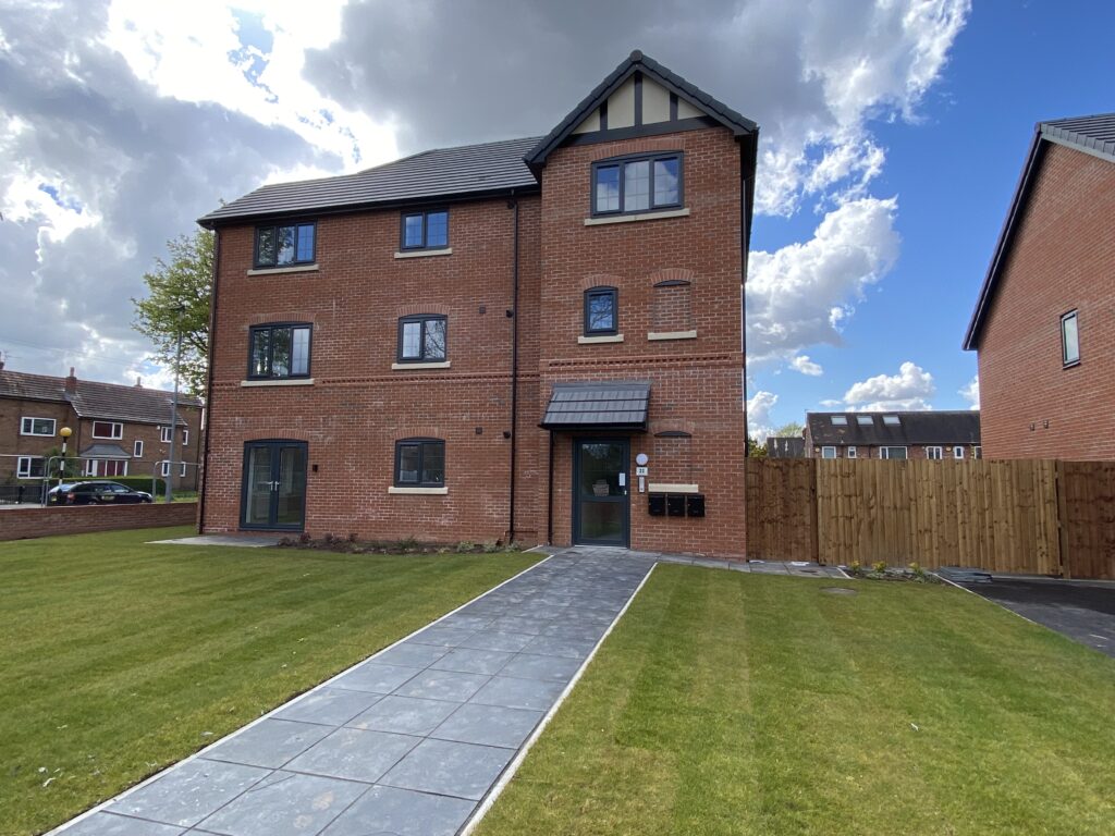 A modern three-story red-brick apartment building with a gray tiled walkway leading to the entrance, surrounded by a neatly mowed green lawn and a wooden fence, under a partly cloudy sky.