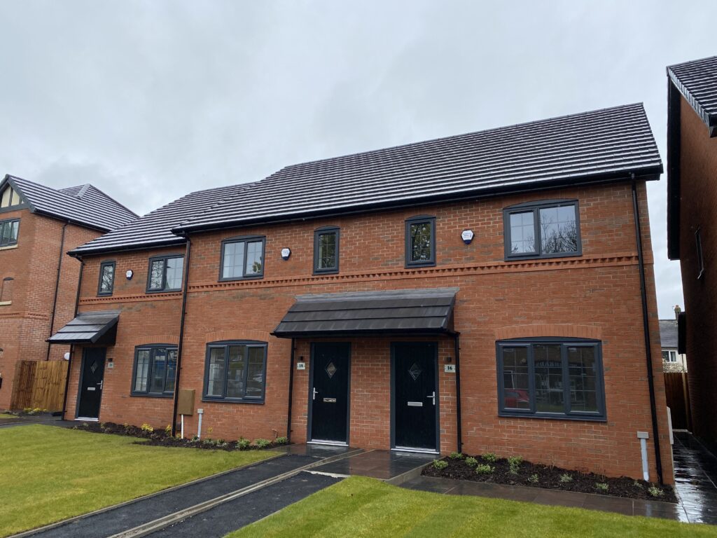 A modern, two-story red brick duplex with dark doors and windows, a tiled roof, and small front lawns on a cloudy day. A path leads to each entrance, and neighboring houses are visible on either side.
