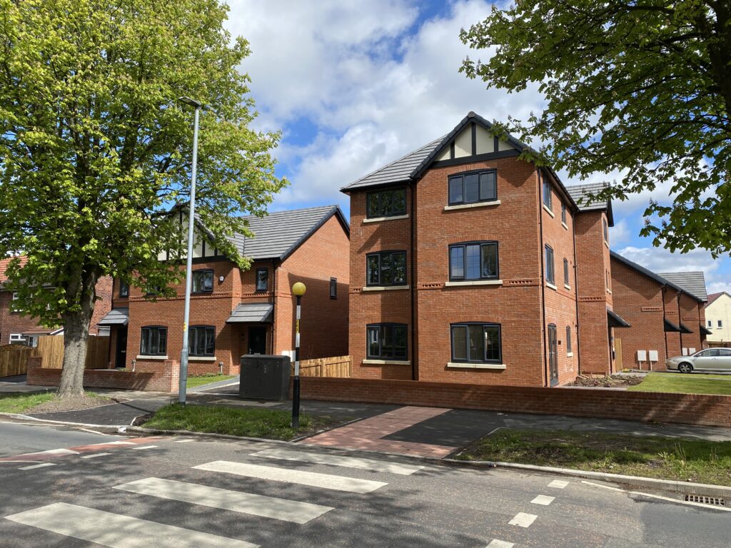 Newly built red-brick houses with black roofs are situated beside a tree-lined street with a zebra crossing, under a partly cloudy sky. Grass lawns and paved walkways surround the homes.