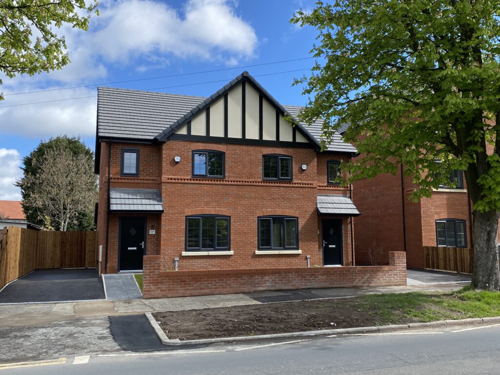A modern red-brick semi-detached house with two black front doors, a small front yard, and a tree on the right side, on a sunny day with blue sky and clouds.