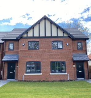 A modern semi-detached red brick house with a gabled roof, black trim, two front doors, and a well-kept green lawn under a partly cloudy sky.