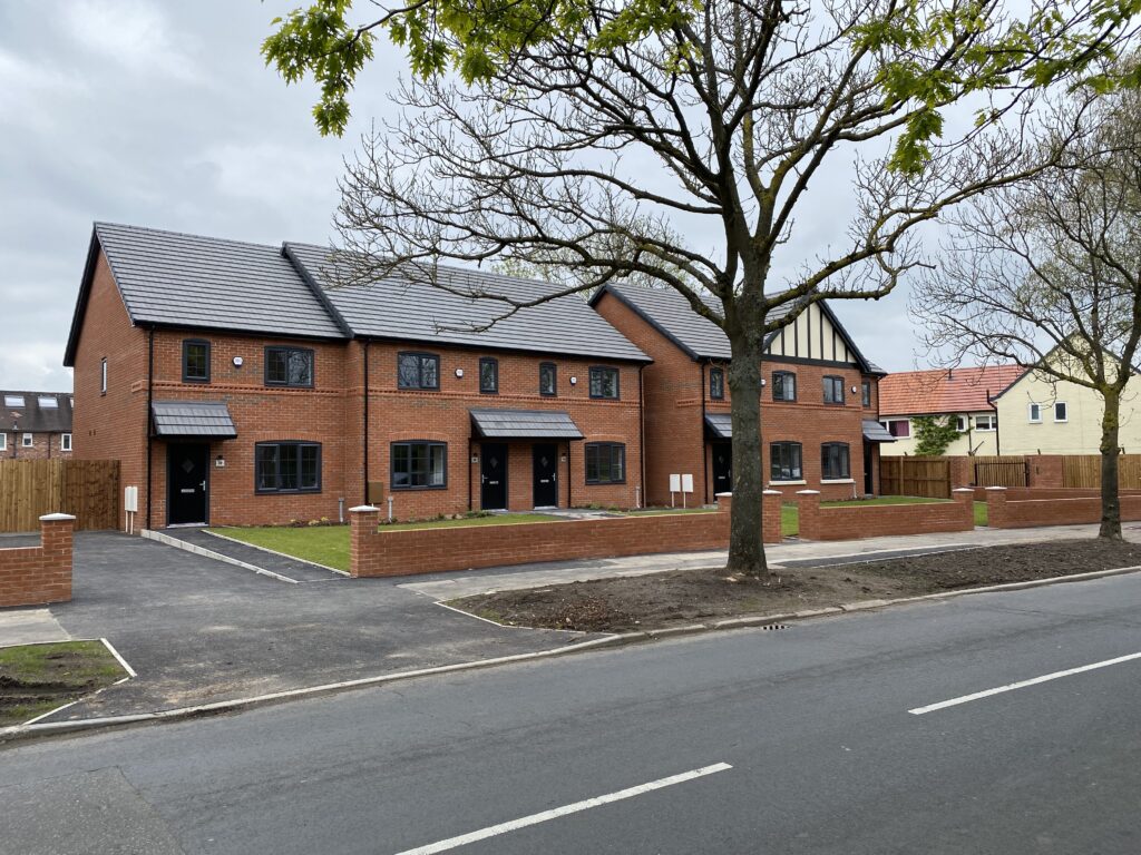 A row of modern red-brick townhouses with dark windows and doors, separated by small lawns and low brick walls, stands along a quiet street with leafless trees and overcast skies.
