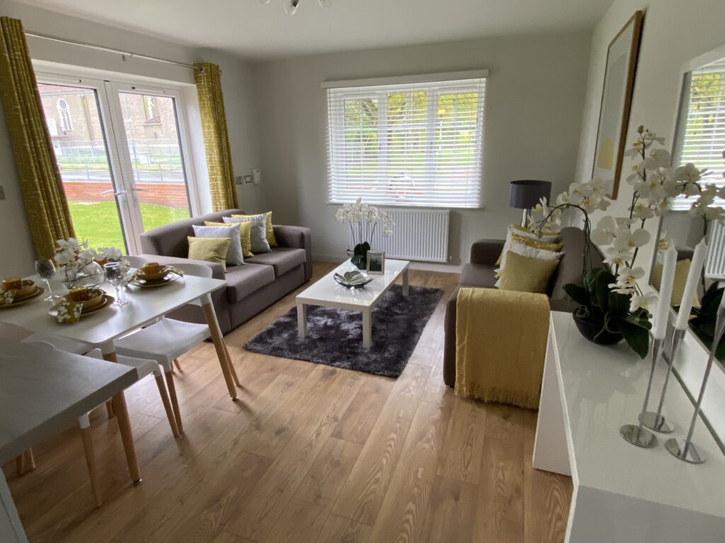 A modern, bright living and dining area with light wood floors, gray sofas, yellow accents, and white furniture. Large windows and glass doors let in natural light, overlooking a green lawn.
