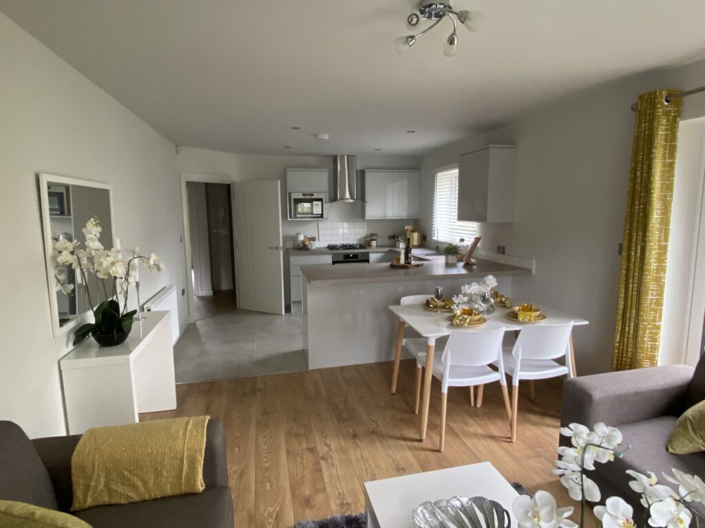 Modern open-plan kitchen and dining area with white cabinets, wood flooring, a dining table set for four, brown sofas, yellow accents, and decorative flowers, with natural light streaming in through a window.