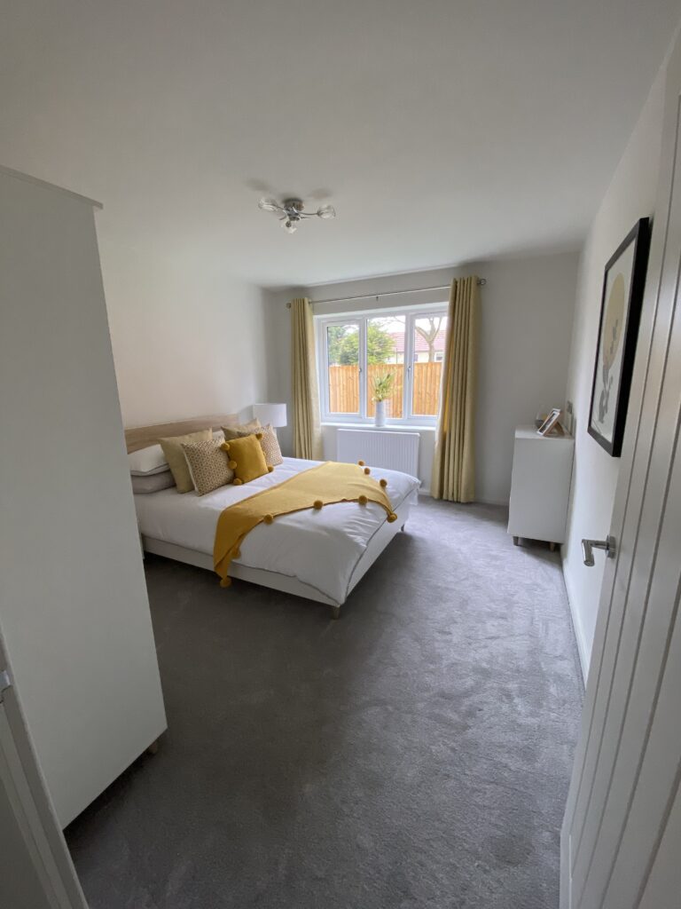 A bright bedroom with light gray carpet, a double bed with white bedding and yellow accents, large window with yellow curtains, and white furniture. Natural light fills the room.