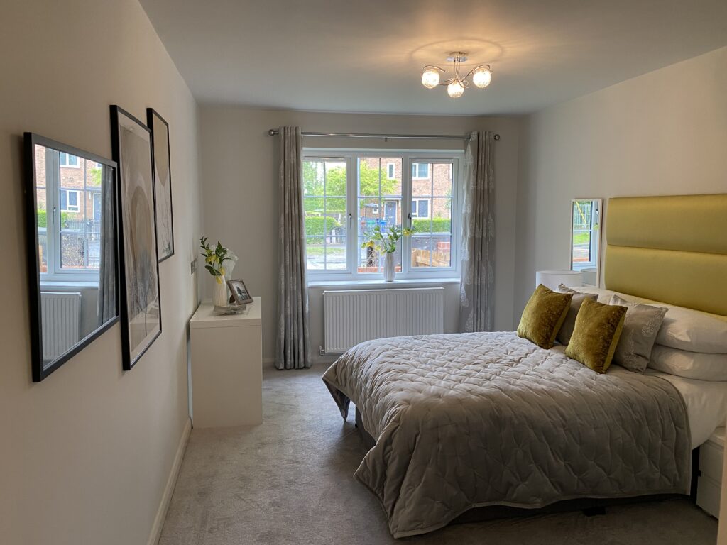 A modern bedroom with a large window, double bed with a yellow headboard, gray bedding, and yellow pillows. There are wall art pieces, a white dresser with a vase and photo frame, and light gray curtains.
