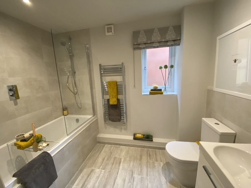 Modern bathroom with a bathtub and shower, towel rack with yellow towels, window with grey patterned blind, toilet, sink, and decorative items in yellow and green accents. Light grey tiles and flooring throughout.