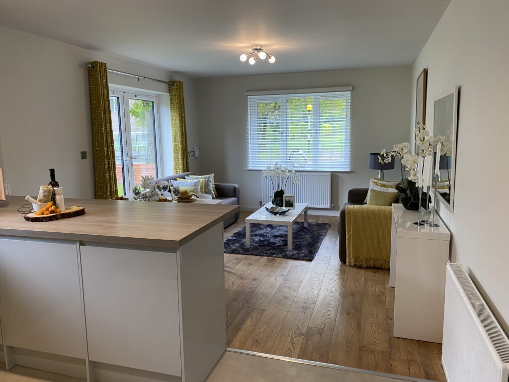 Modern open-plan living room with wood flooring, gray sofa, yellow accents, large windows with blinds, white walls, kitchen island in foreground, and decorative plants and flowers throughout.