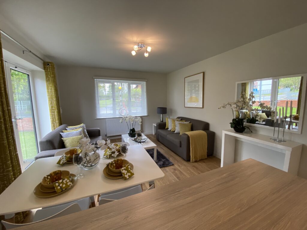 A modern, bright living-dining room with two grey sofas, yellow accents, a set dining table for four, large windows, and decorative plants. Light wood floors and white walls create an airy atmosphere.