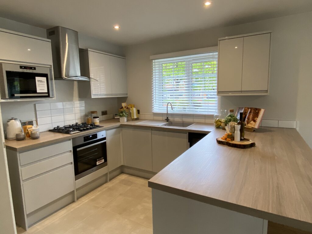 Modern kitchen with white cabinets, stainless steel appliances, gas stove, microwave, and a sink under a window with blinds. Decor includes plants, a breadboard, and neatly arranged kitchen items.