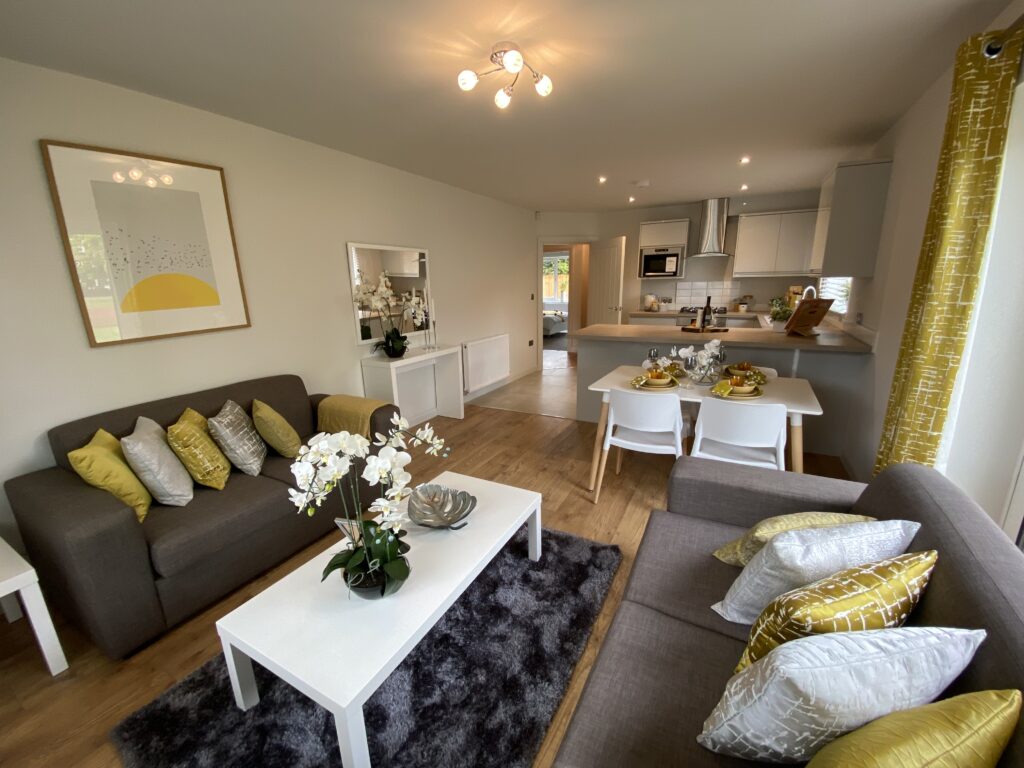 Modern open-plan living and dining area with two gray sofas, yellow and silver cushions, white tables, a rug, wall art, and a kitchen in the background. Large windows with yellow curtains brighten the space.