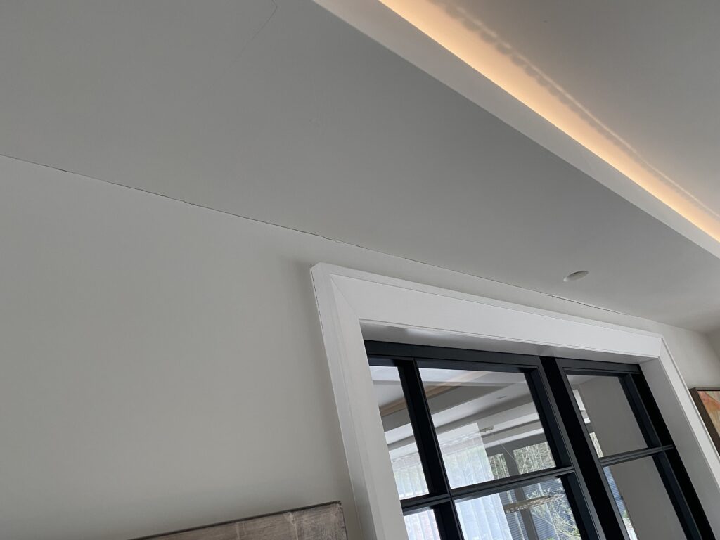 A white ceiling with a visible crack running parallel to the wall above a window with black-framed glass panes; recessed lighting is installed near the ceiling.