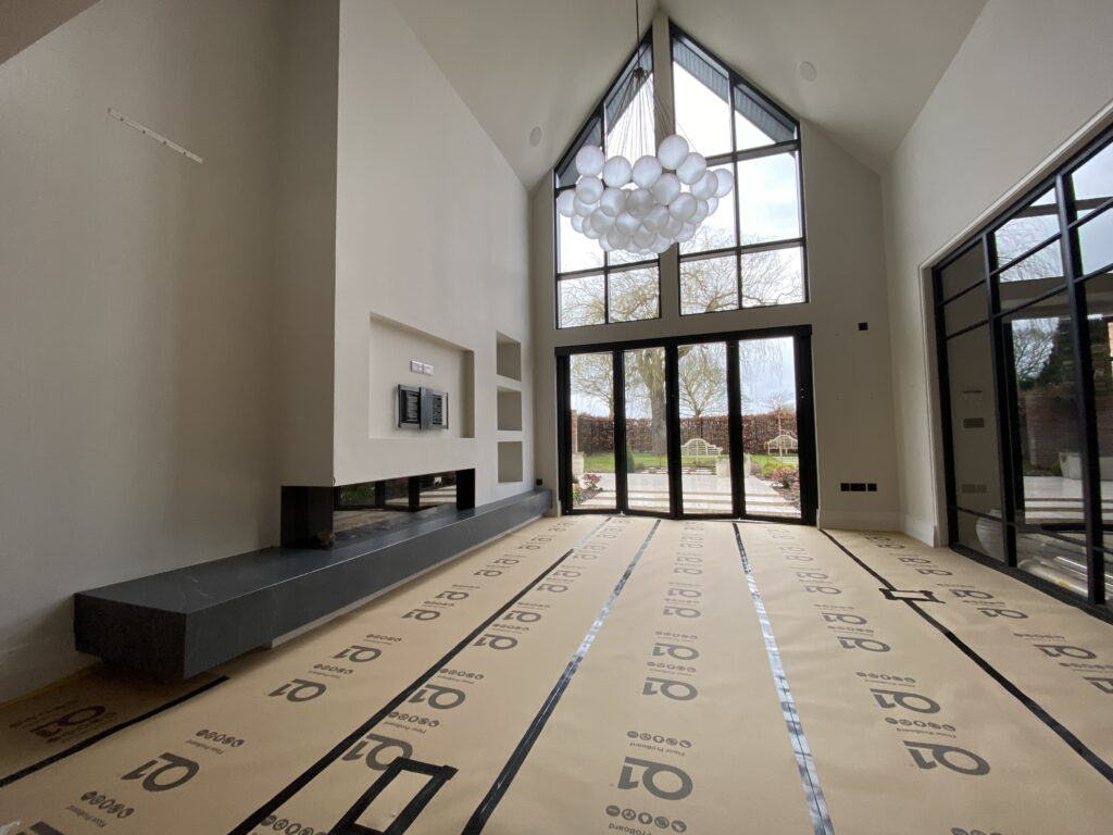 A modern, unfurnished living room with tall, vaulted ceilings, large floor-to-ceiling windows, a contemporary fireplace, and a cluster of round pendant lights. The floor is covered with protective paper.