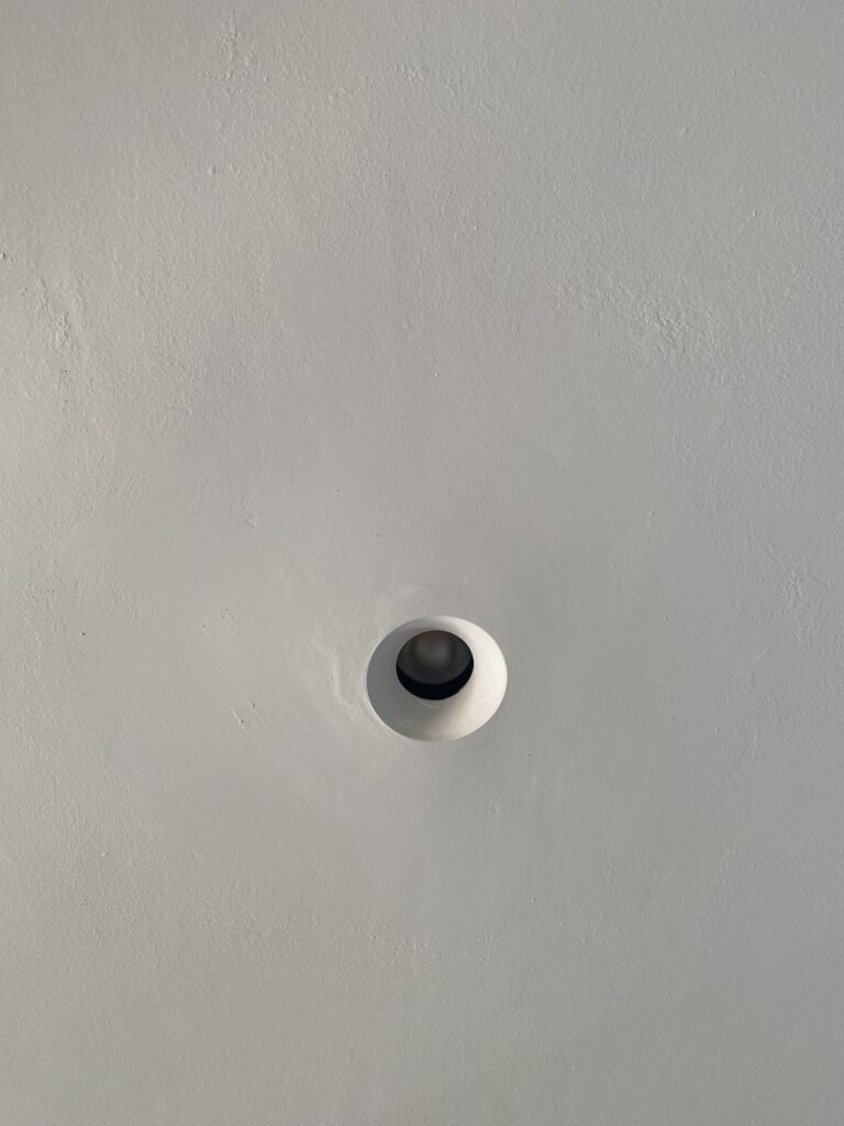 A close-up of a plain white or light gray ceiling with a single, round recessed light fixture in the center. The fixture appears empty, with no visible bulb inside.