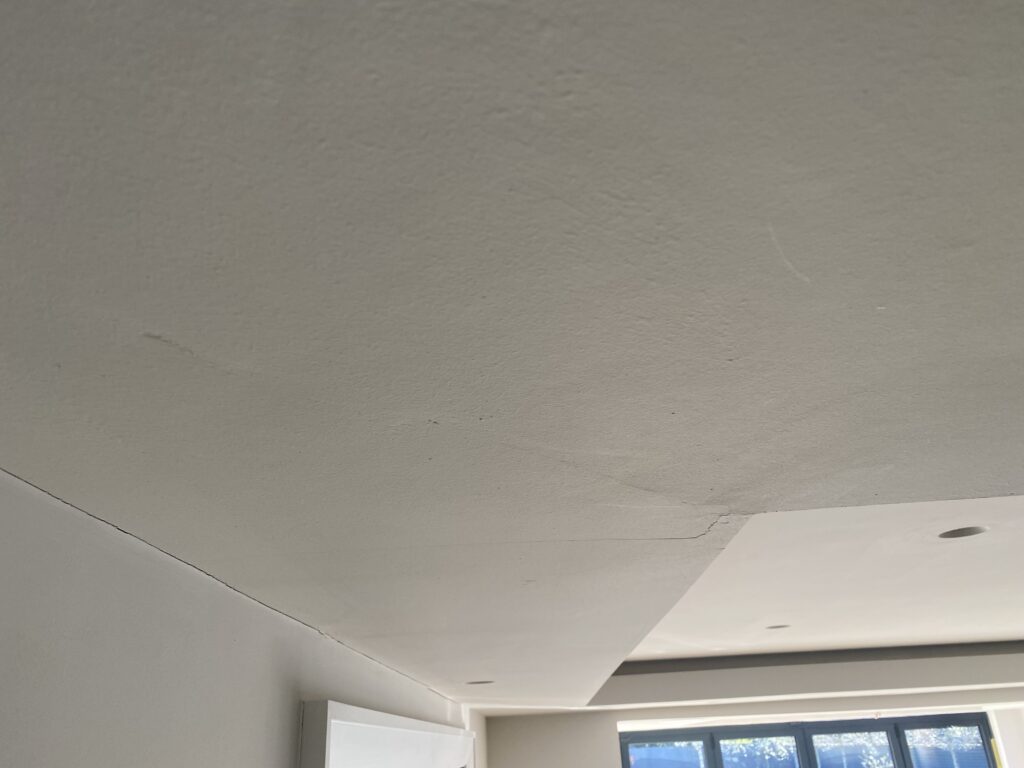 A ceiling with visible cracks and seams, showing an uneven surface and slight discoloration, with recessed lighting and a window letting in daylight in the background.