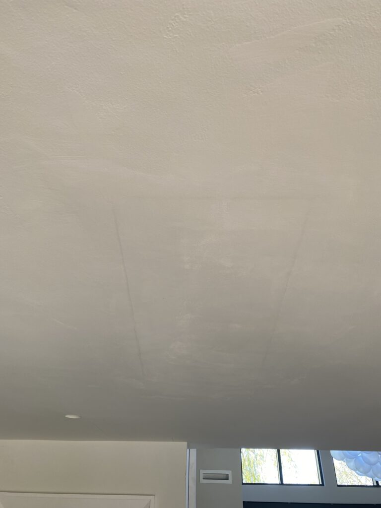 A ceiling with faint, visible lines and subtle discoloration; part of a wall, a vent, a recessed light, and a window with trees visible through it are also shown at the bottom of the image.