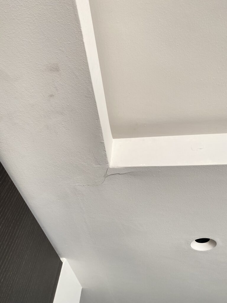 A white ceiling with a recessed tray design shows a visible crack where two sections meet. There is a round recessed light fixture nearby and some faint discoloration on the ceiling.