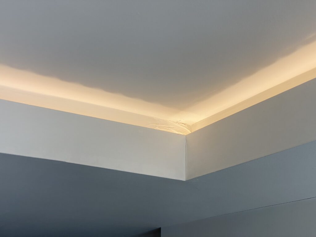 A ceiling corner with recessed lighting casting a soft glow along the edge, highlighting a slightly uneven or cracked area where the walls and ceiling meet.