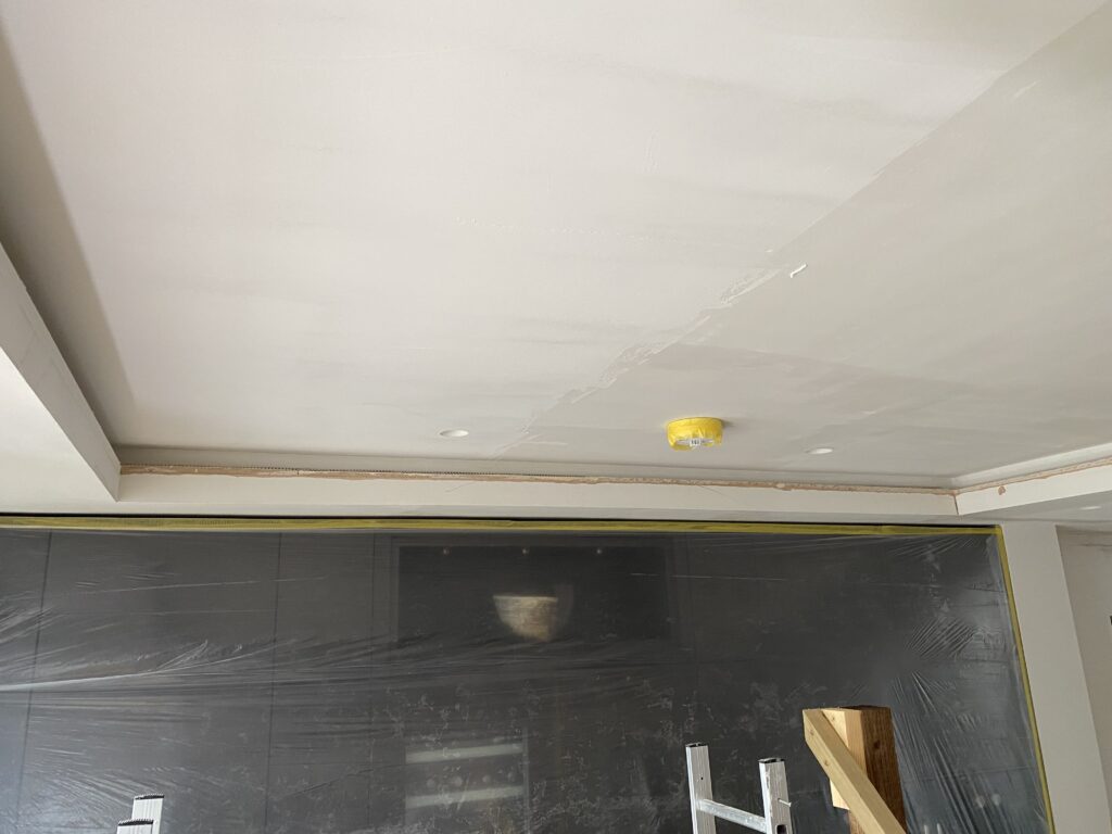 A room under renovation with a freshly plastered ceiling, covered light fixtures, and plastic sheeting protecting the walls and cabinets. A wooden ladder is partially visible in the foreground.