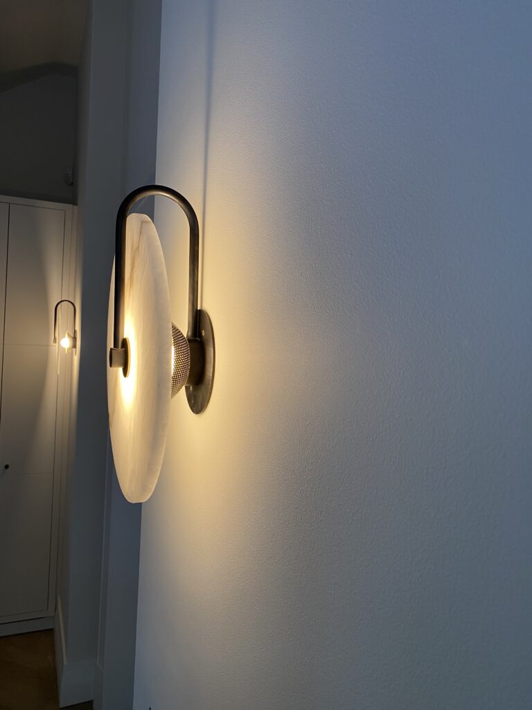 Modern wall sconce with a round white shade emits warm light, casting a soft glow and shadow on a light-colored wall; a similar sconce is visible further down the hallway.