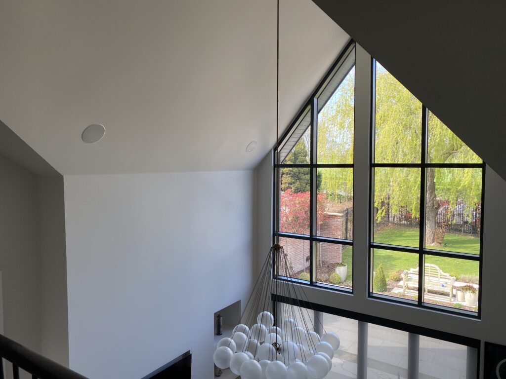 A modern living room with tall, slanted ceilings, large black-framed windows, a cluster of white pendant lights, and a view of a green backyard with trees and outdoor furniture.