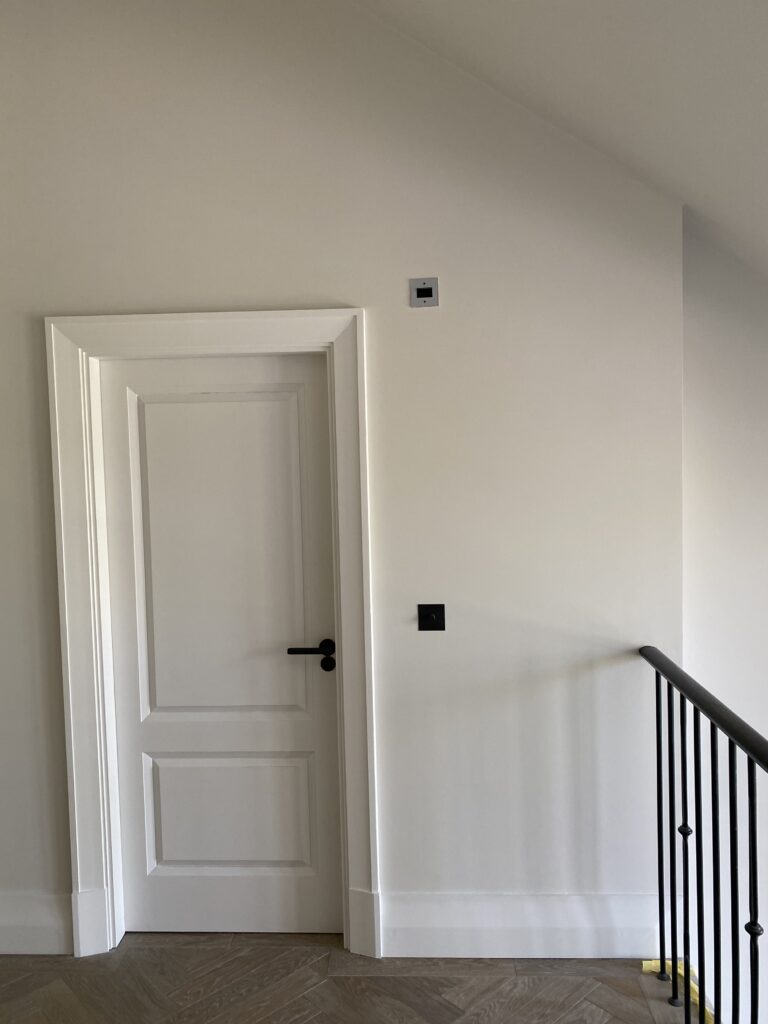 A white interior door with a black handle is set in a light-colored wall. There are two small square fixtures above and beside the door, and a black railing is visible on the right side. Wooden flooring is at the bottom.