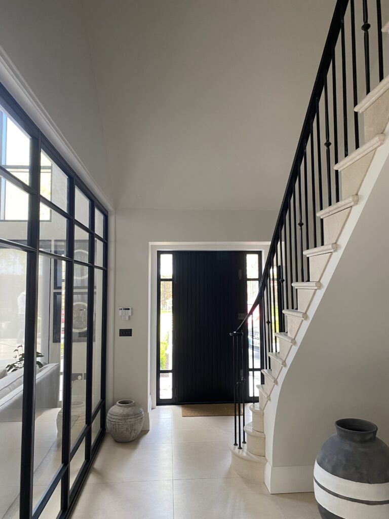 Bright modern entryway with a black front door, large glass side window, a white staircase with black railings, light tiled floor, and two decorative vases placed near the stairs and the window.